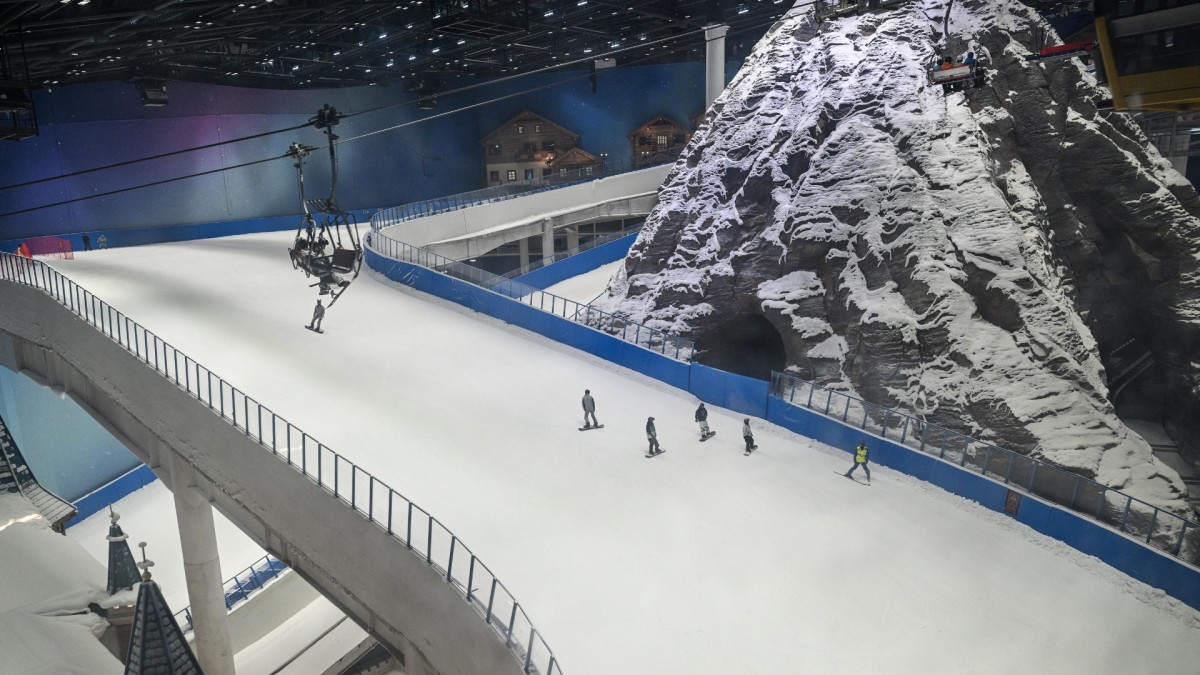 A man dressed in a yellow jacket is lying injured on the snow-covered slope at Huafa Ice and Snow World indoor ski resort, while several other skiers are walking towards him and a ski lift is visible above.